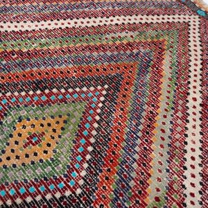 May include: Close-up of a colourful, patterned rug with a geometric design. The rug features a diamond pattern in the centre, surrounded by stripes of red, green, blue, and orange. The rug's edges have a fringed border.