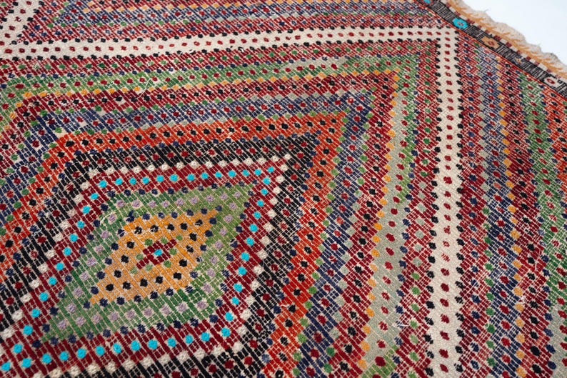 May include: Close-up of a colourful, patterned rug with a geometric design. The rug features a diamond pattern in the centre, surrounded by stripes of red, green, blue, and orange. The rug's edges have a fringed border.