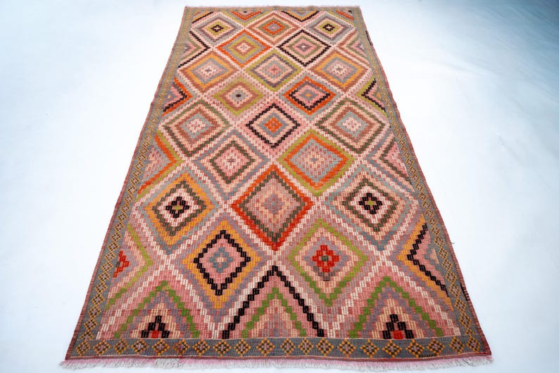 May include: A rectangular rug with a geometric diamond pattern. The rug features a variety of colors, including pink, orange, green, brown, and gray. The diamond pattern is repeated across the rug's surface, creating a visually appealing design. The rug has a fringed edge.