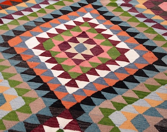 Può includere: Un tappeto geometrico colorato con un motivo a rombi. Il tappeto presenta triangoli nelle tonalità di arancione, blu, verde, rosa, bianco e nero. Il tappeto è probabilmente realizzato in tessuto intrecciato.