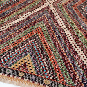 May include: Close-up of a colourful, patterned rug with a geometric design. The rug features a variety of colours, including red, green, blue, and beige, arranged in diagonal stripes and diamond shapes. The rug has a fringed edge.