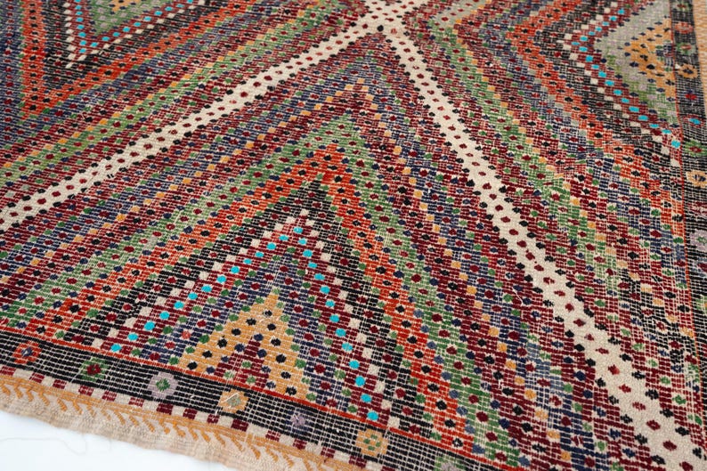 May include: Close-up of a colourful, patterned rug with a geometric design. The rug features a variety of colours, including red, green, blue, and beige, arranged in diagonal stripes and diamond shapes. The rug has a fringed edge.