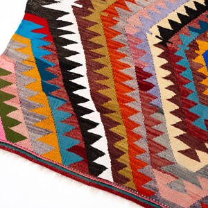 K&ouml;nnte beinhalten: Ein farbenfroher, geometrisch gemusterter Teppich mit einem rautenf&ouml;rmigen zentralen Design. Der Teppich weist eine Vielzahl von Farben auf, darunter Rot, Blau, Gelb, Orange und Wei&szlig;. Die R&auml;nder des Teppichs sind gefranst.