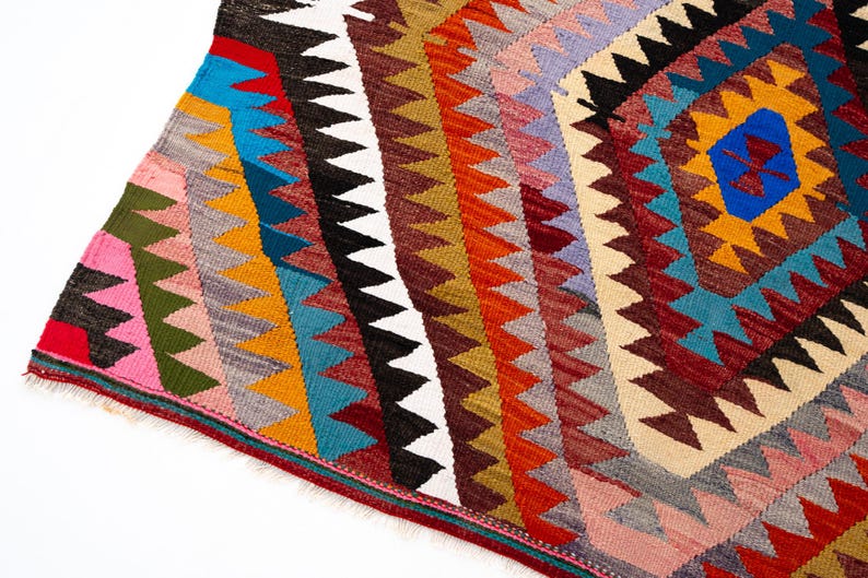 K&ouml;nnte beinhalten: Ein farbenfroher, geometrisch gemusterter Teppich mit einem rautenf&ouml;rmigen zentralen Design. Der Teppich weist eine Vielzahl von Farben auf, darunter Rot, Blau, Gelb, Orange und Wei&szlig;. Die R&auml;nder des Teppichs sind gefranst.