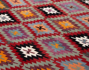 Può includere: Un tappeto fantasia con un design geometrico in una varietà di colori. Il tappeto presenta un motivo ripetuto di forme a diamante in rosso, nero, bianco, arancione, viola e blu. Il tappeto ha un aspetto strutturato.
