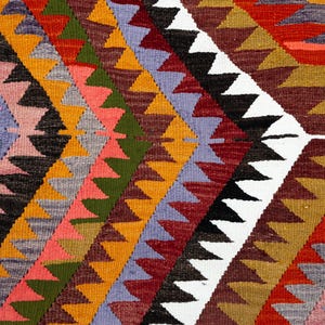 K&ouml;nnte beinhalten: Nahaufnahme eines gewebten Textils mit geometrischem Muster. Das Design zeigt Reihen farbiger Dreiecke in Orange-, Rot-, Gr&uuml;n-, Rosa- und Lilat&ouml;nen vor einem wei&szlig;en Hintergrund. Das Textil scheint ein Teppich oder Wandbehang zu sein.