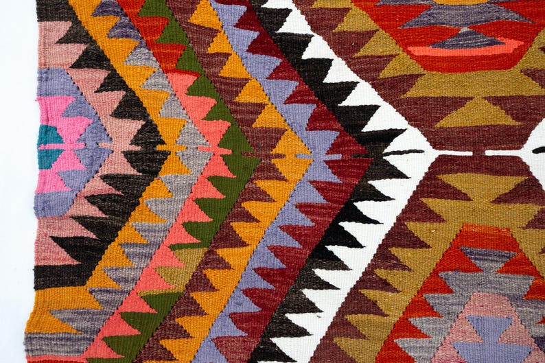 K&ouml;nnte beinhalten: Nahaufnahme eines gewebten Textils mit geometrischem Muster. Das Design zeigt Reihen farbiger Dreiecke in Orange-, Rot-, Gr&uuml;n-, Rosa- und Lilat&ouml;nen vor einem wei&szlig;en Hintergrund. Das Textil scheint ein Teppich oder Wandbehang zu sein.