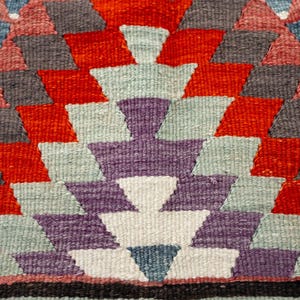 Puede incluir: Primer plano de un textil tejido con un patr&oacute;n geom&eacute;trico. El dise&ntilde;o presenta formas de diamante en rojo, gris, morado y azul, dispuestas en un patr&oacute;n sim&eacute;trico. El fondo es una mezcla de colores, creando una apariencia texturizada.