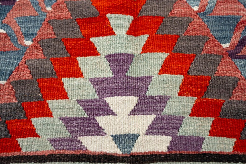 Puede incluir: Primer plano de un textil tejido con un patr&oacute;n geom&eacute;trico. El dise&ntilde;o presenta formas de diamante en rojo, gris, morado y azul, dispuestas en un patr&oacute;n sim&eacute;trico. El fondo es una mezcla de colores, creando una apariencia texturizada.