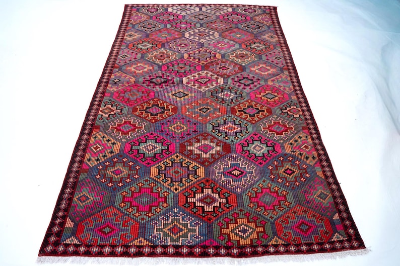 May include: A rectangular area rug featuring a repeating geometric pattern of interconnected hexagons and diamonds in vibrant shades of hot pink, deep purple, orange, and teal, finished with a dark maroon border and short white fringe at both ends.