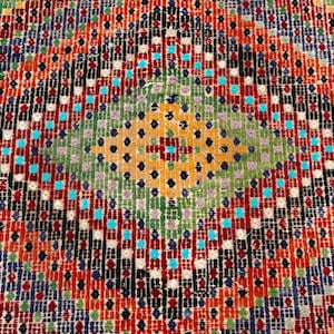 May include: Close-up of a colourful, patterned rug. The design features a central diamond shape with a green and yellow centre, surrounded by red, orange, black, and blue geometric patterns. The rug has a textured appearance.