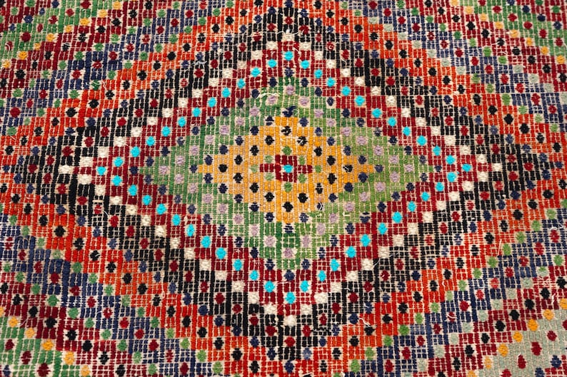 May include: Close-up of a colourful, patterned rug. The design features a central diamond shape with a green and yellow centre, surrounded by red, orange, black, and blue geometric patterns. The rug has a textured appearance.