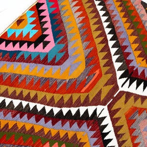 K&ouml;nnte beinhalten: Ein farbenfroher, geometrischer Teppich mit einem sich wiederholenden Dreiecksmuster in Rot-, Orange-, Gelb-, Braun-, Rosa-, Blau- und Schwarzt&ouml;nen. Der Teppich hat eine strukturierte Optik und einen wei&szlig;en Rand.