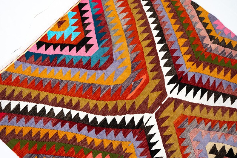 K&ouml;nnte beinhalten: Ein farbenfroher, geometrischer Teppich mit einem sich wiederholenden Dreiecksmuster in Rot-, Orange-, Gelb-, Braun-, Rosa-, Blau- und Schwarzt&ouml;nen. Der Teppich hat eine strukturierte Optik und einen wei&szlig;en Rand.
