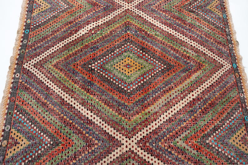 May include: A vibrant, geometric patterned rug with a diamond design. The rug features a variety of colours including red, green, orange, and white. The rug has a fringed edge.