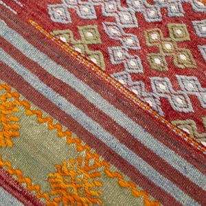 May include: Close-up of a section of a woven rug with a geometric pattern. The rug features a red background with blue and white diamond shapes. There are also stripes of red, blue, and green with orange embroidered details.