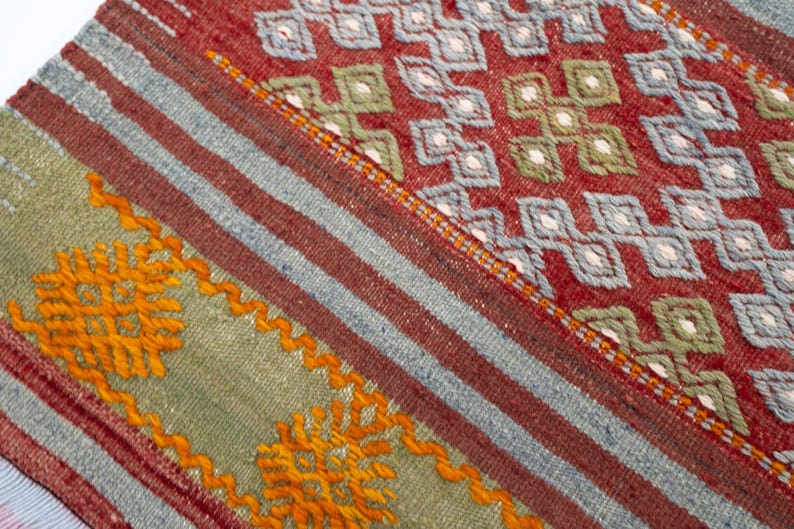 May include: Close-up of a section of a woven rug with a geometric pattern. The rug features a red background with blue and white diamond shapes. There are also stripes of red, blue, and green with orange embroidered details.