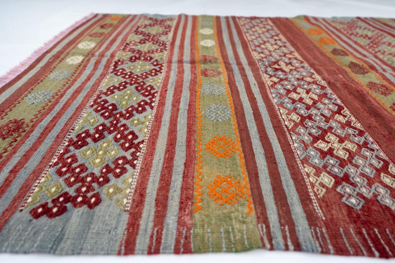 May include: A rectangular rug with a striped pattern in shades of red, gray, green, and orange. The rug features geometric designs and a fringed edge. The rug is made of woven fabric.