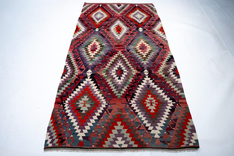 Puede incluir: Una alfombra rectangular tejida a mano con un patr&oacute;n geom&eacute;trico de diamantes. La alfombra presenta una vibrante paleta de colores de rojo, morado, verde y blanco. El dise&ntilde;o es sim&eacute;trico, con formas de diamantes repetidas. Esta alfombra es adecuada para la decoraci&oacute;n del hogar.