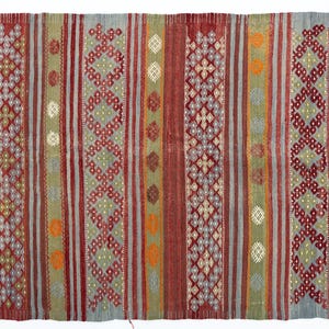 May include: A rectangular, woven rug with a striped pattern in shades of red, green, gray, and orange. The rug features geometric diamond and dot designs. The edges have a pink fringe.