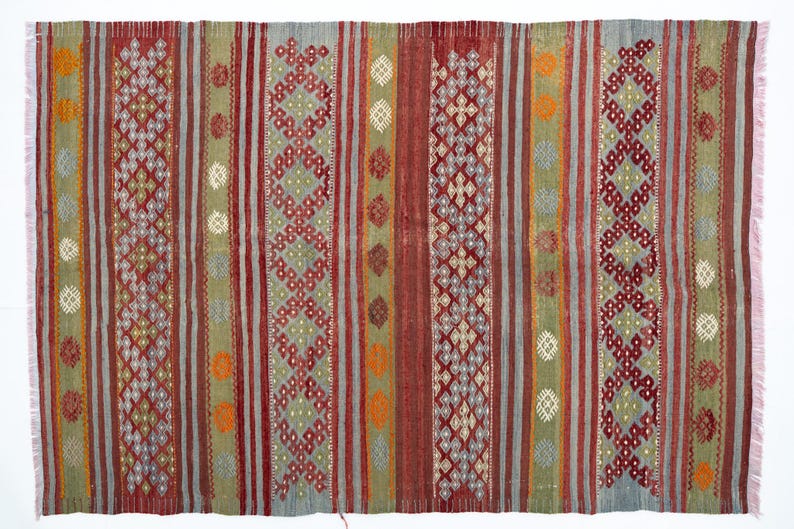 May include: A rectangular, woven rug with a striped pattern in shades of red, green, gray, and orange. The rug features geometric diamond and dot designs. The edges have a pink fringe.