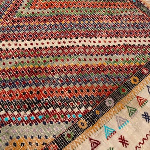 May include: Close-up of a patterned rug with a geometric design. The rug features red, orange, blue, green, and cream colours. The edges have a decorative border with fringe.
