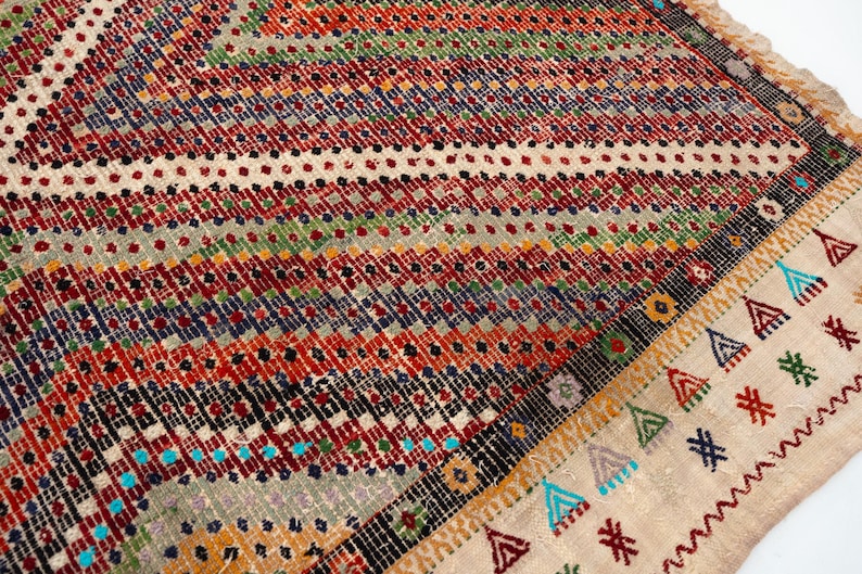 May include: Close-up of a patterned rug with a geometric design. The rug features red, orange, blue, green, and cream colours. The edges have a decorative border with fringe.