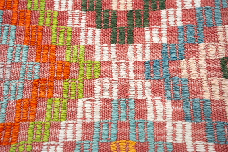 May include: Close-up of a woven textile with a colorful, geometric pattern. The design features alternating blocks of red, white, green, orange, and blue. The texture appears soft and the weave is tight.