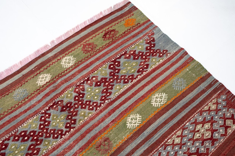 May include: A section of a woven rug with a geometric pattern. The rug features a combination of colors, including red, green, gray, and orange. The design includes stripes and diamond shapes. The rug has a pink fringe.