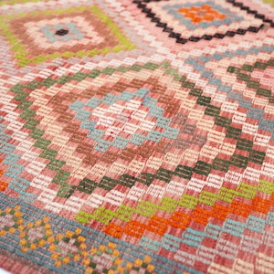 May include: Close-up of a woven rug with a geometric pattern. The rug features a repeating diamond and rectangular design in shades of pink, orange, green, blue, and brown. The texture of the rug is visible.