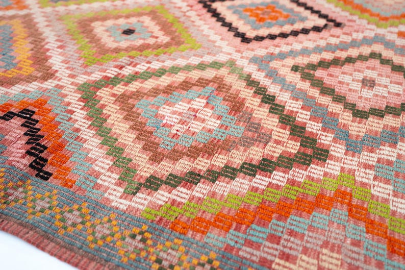 May include: Close-up of a woven rug with a geometric pattern. The rug features a repeating diamond and rectangular design in shades of pink, orange, green, blue, and brown. The texture of the rug is visible.