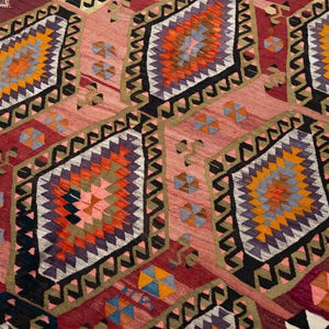 May include: A close-up of a handmade rug with a geometric pattern. The rug features diamond shapes in shades of red, orange, purple, and yellow, with a black and gold border. The background is a lighter shade of red.