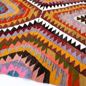 K&ouml;nnte beinhalten: Ein farbenfroher, geometrisch gemusterter Teppich mit einer Vielzahl von Farben, darunter Orange, Gelb, Rosa und Gr&uuml;n. Der Teppich weist ein sich wiederholendes dreieckiges Design mit wei&szlig;en Akzenten auf. Der Teppich besteht aus einem gewebten Material.