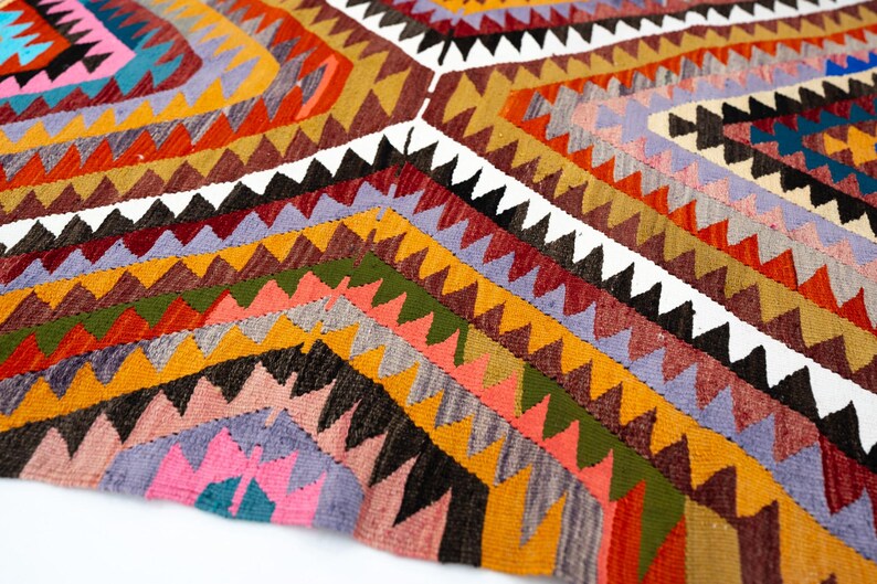 K&ouml;nnte beinhalten: Ein farbenfroher, geometrisch gemusterter Teppich mit einer Vielzahl von Farben, darunter Orange, Gelb, Rosa und Gr&uuml;n. Der Teppich weist ein sich wiederholendes dreieckiges Design mit wei&szlig;en Akzenten auf. Der Teppich besteht aus einem gewebten Material.