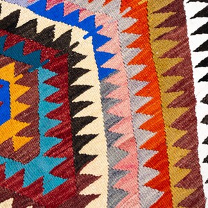 K&ouml;nnte beinhalten: Nahaufnahme eines farbenfrohen, geometrisch gemusterten Teppichs. Der Teppich hat eine zentrale Diamantenform mit blauem Zentrum und gelbem Rand. Um den Diamanten herum befinden sich Dreiecke in verschiedenen Farben, darunter Rot, Blau und Schwarz.