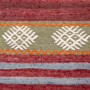 May include: Close-up of a woven textile with a geometric pattern. The rug features a combination of red, green, and blue stripes, with white and orange accents. The central design includes two white diamond-shaped motifs on a green background.