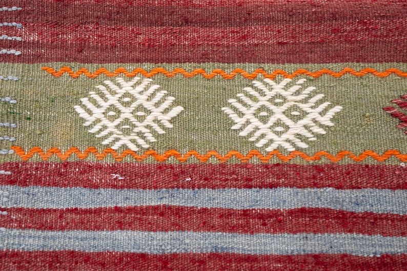 May include: Close-up of a woven textile with a geometric pattern. The rug features a combination of red, green, and blue stripes, with white and orange accents. The central design includes two white diamond-shaped motifs on a green background.