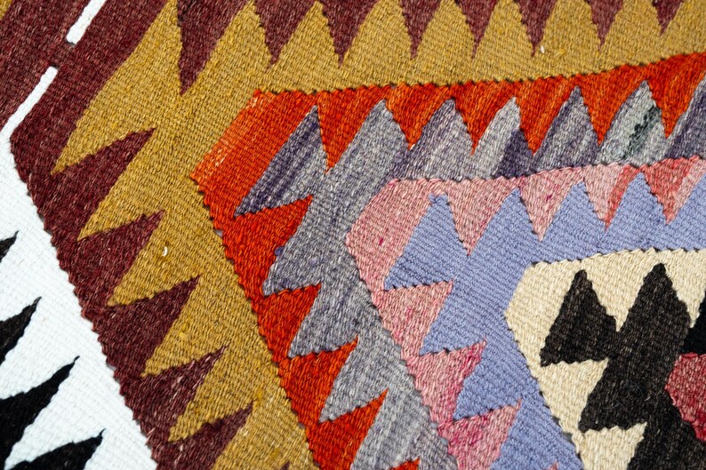 K&ouml;nnte beinhalten: Nahaufnahme eines gewebten Textils mit geometrischem Muster. Das Design zeigt sich wiederholende Dreiecksformen in Gold-, Rot-, Grau-, Rosa-, Blau-, Schwarz- und Wei&szlig;t&ouml;nen. Die Textur des Stoffes ist sichtbar.
