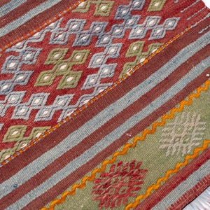 May include: Close-up of a woven rug with a geometric pattern. The rug features a red background with blue and white diamond shapes, green diamond shapes, and red and orange accents. The rug has a striped design with a green border.