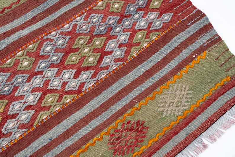 May include: Close-up of a woven rug with a geometric pattern. The rug features a red background with blue and white diamond shapes, green diamond shapes, and red and orange accents. The rug has a striped design with a green border.