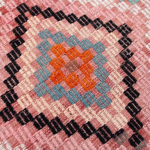 May include: Close-up of a woven textile with a geometric pattern. The design features a central square with orange and blue accents, surrounded by a black border. The background is a mix of pink, white, and green.