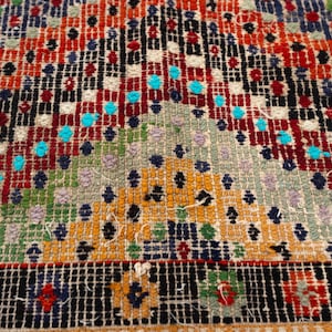 May include: Close-up of a patterned rug with a vibrant colour scheme. The rug displays a geometric design with red, orange, blue, green, and yellow hues. The pattern is detailed, featuring small squares and shapes.