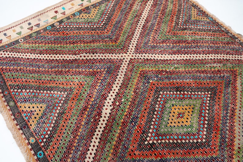 May include: A vibrant, geometric patterned rug with a diamond design. The rug has a cream-coloured border with small, colourful embroidered details. The main body of the rug showcases a series of diamond shapes in shades of red, green, orange, and blue.