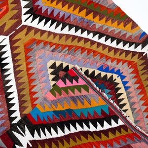 K&ouml;nnte beinhalten: Ein farbenfroher, geometrisch gemusterter Teppich mit einer Vielzahl von Farben, darunter Rot, Orange, Blau und Wei&szlig;. Der Teppich weist ein sich wiederholendes Rautenmuster mit Zickzackkanten auf. Der Teppich besteht aus gewebtem Stoff.
