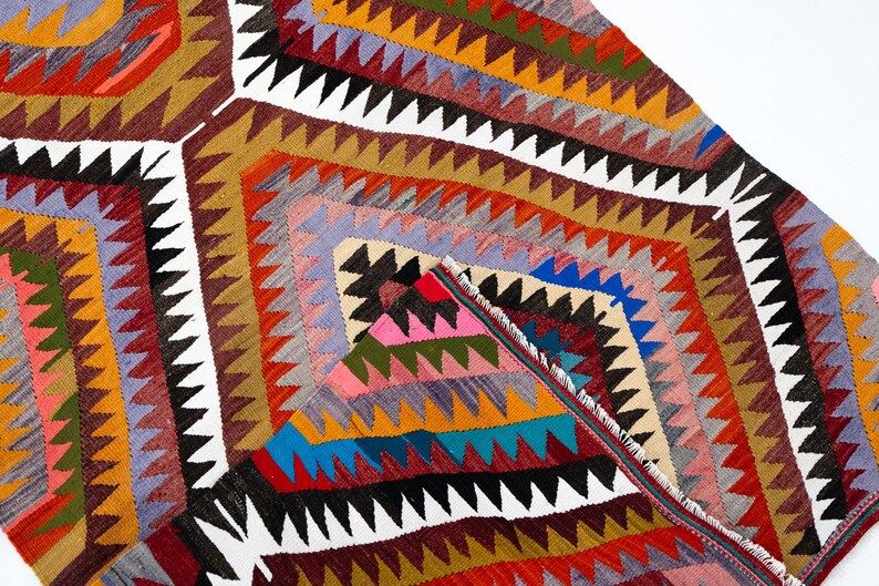 K&ouml;nnte beinhalten: Ein farbenfroher, geometrisch gemusterter Teppich mit einer Vielzahl von Farben, darunter Rot, Orange, Blau und Wei&szlig;. Der Teppich weist ein sich wiederholendes Rautenmuster mit Zickzackkanten auf. Der Teppich besteht aus gewebtem Stoff.