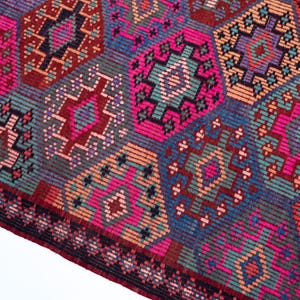May include: A woven textile rug featuring a repeating geometric pattern of diamonds and hexagons in vibrant shades of magenta, teal, orange, purple, and dark blue. The design includes intricate linear details and a dark border along the edge.