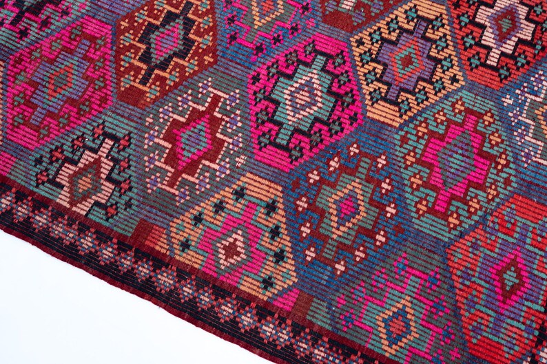 May include: A woven textile rug featuring a repeating geometric pattern of diamonds and hexagons in vibrant shades of magenta, teal, orange, purple, and dark blue. The design includes intricate linear details and a dark border along the edge.