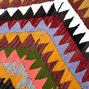 K&ouml;nnte beinhalten: Nahaufnahme eines farbenfrohen gewebten Textils mit geometrischem Muster. Das Design zeigt Reihen von Dreiecken in verschiedenen Farben, darunter Rot, Orange, Gr&uuml;n und Lila. Das Textil hat ein strukturiertes Aussehen.