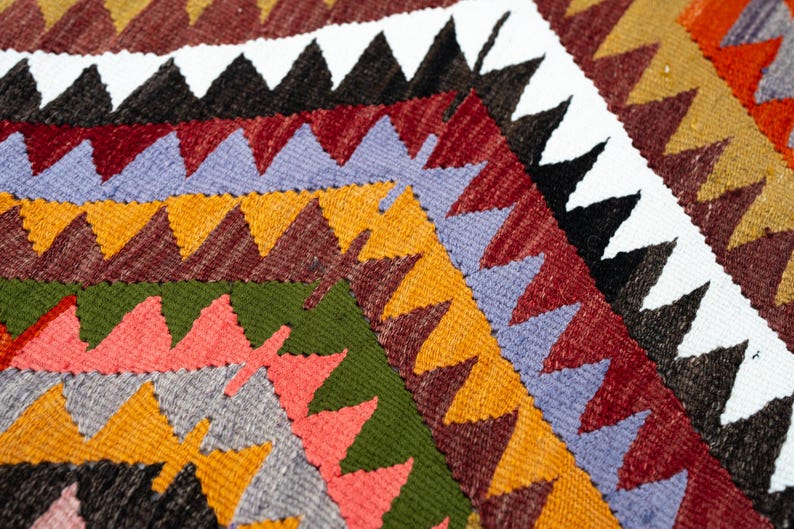 K&ouml;nnte beinhalten: Nahaufnahme eines farbenfrohen gewebten Textils mit geometrischem Muster. Das Design zeigt Reihen von Dreiecken in verschiedenen Farben, darunter Rot, Orange, Gr&uuml;n und Lila. Das Textil hat ein strukturiertes Aussehen.