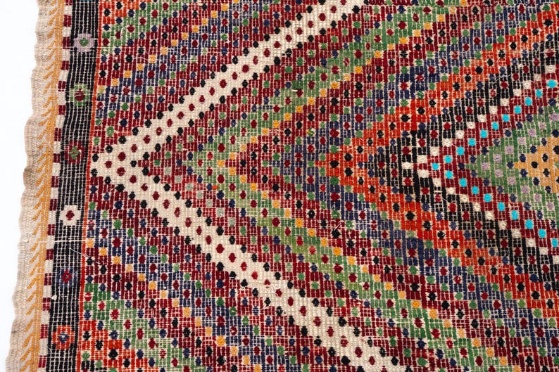 May include: Close-up of a colourful, patterned rug with a geometric design. The rug features a variety of colours including red, green, blue, orange, and white. The edges have a fringed border.